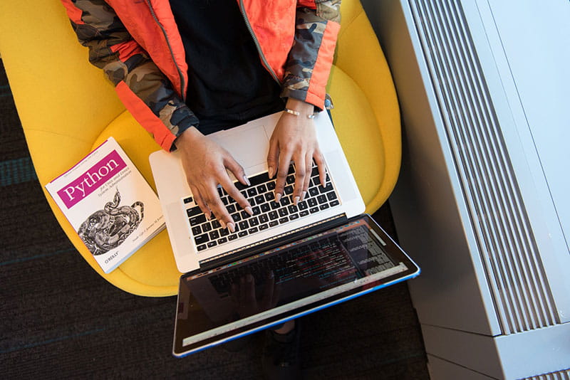 An aerial view of a person typing on a laptop with a Python coding language book on the seat next to them. An aerial view of a person typing on a laptop with a Python coding language book on the seat next to them.